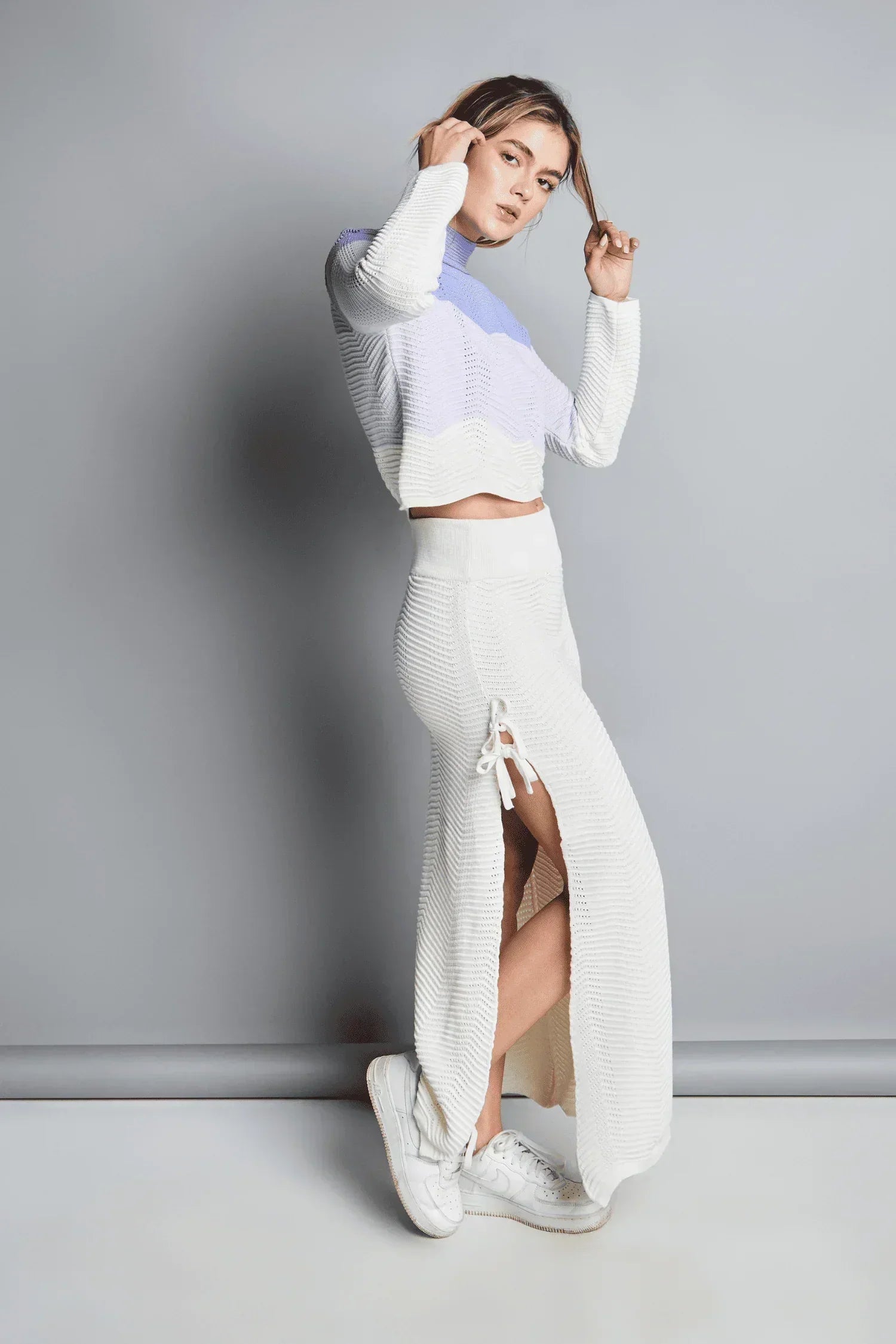 Mujer con conjunto tejido de punto blanco y lila, falda larga con abertura y tenis blancos.